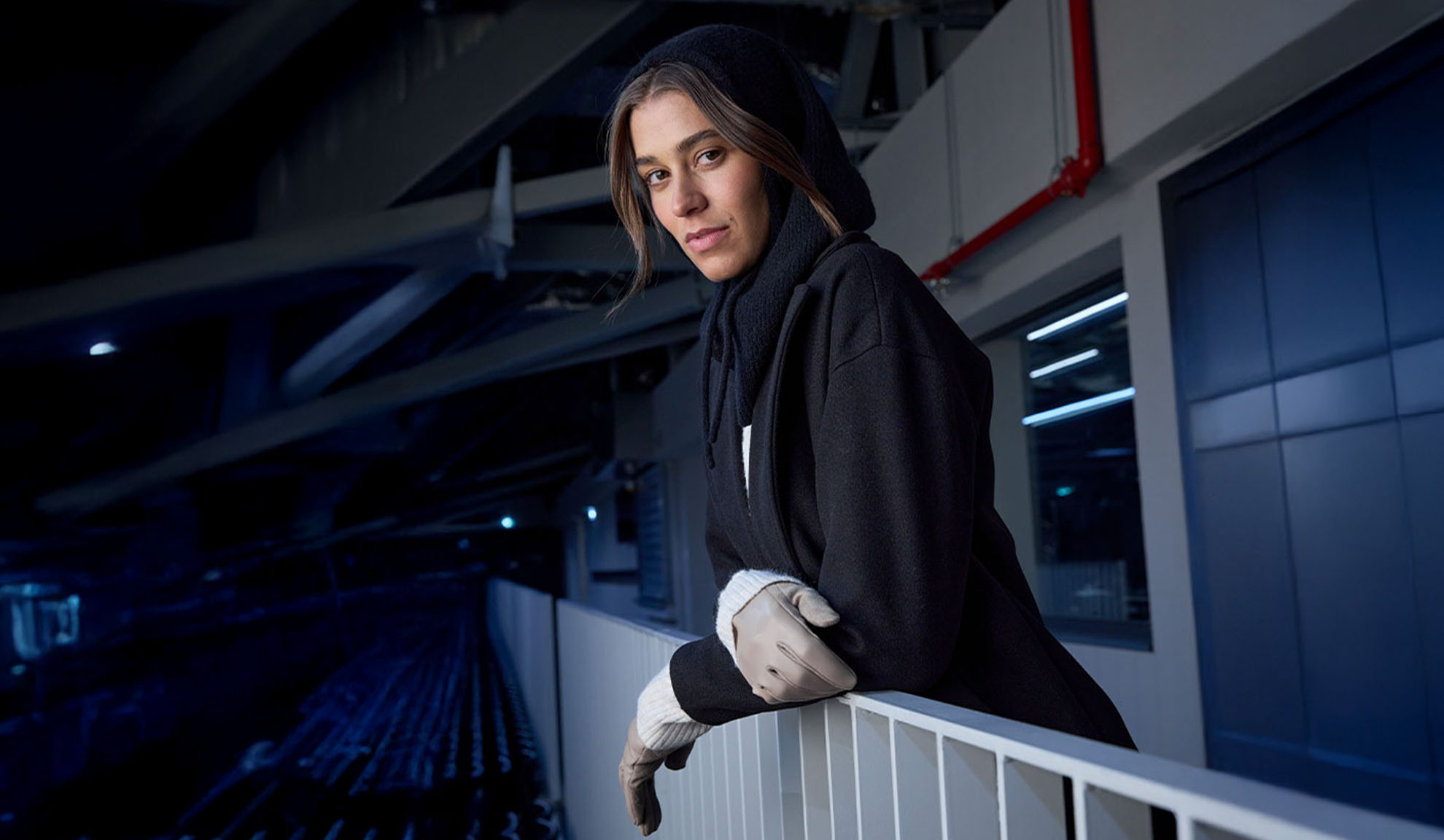 Vrouw in zwarte jas en capuchonsjaal, leunend op een reling in een moderne omgeving.