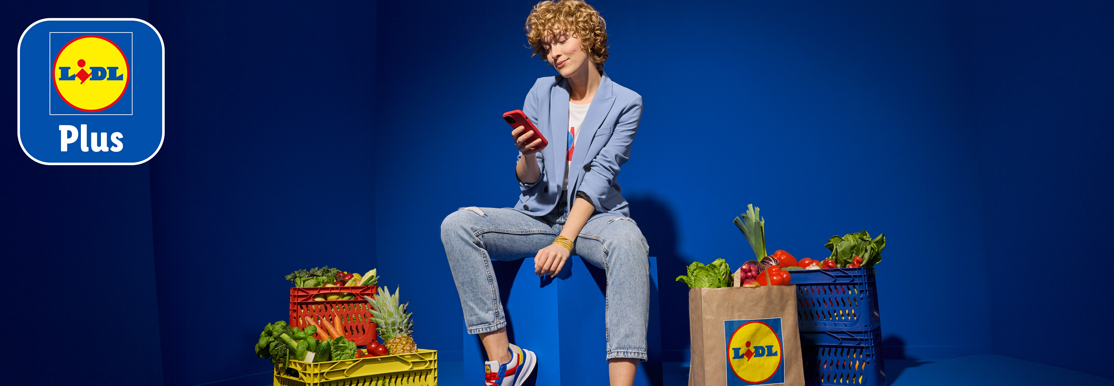 Een vrouw zit op een blauwe kubus en kijkt op haar telefoon, omringd door kratten en tassen vol boodschappen van Lidl.