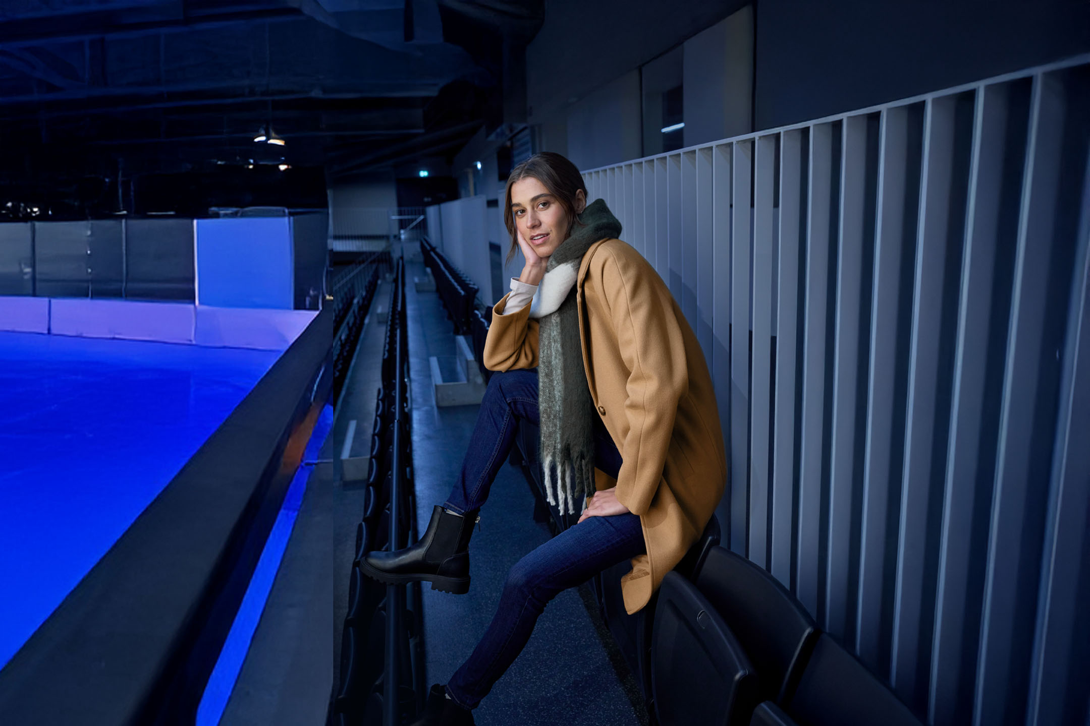 Vrouw in jas en sjaal zit op de tribune van een ijsbaan.