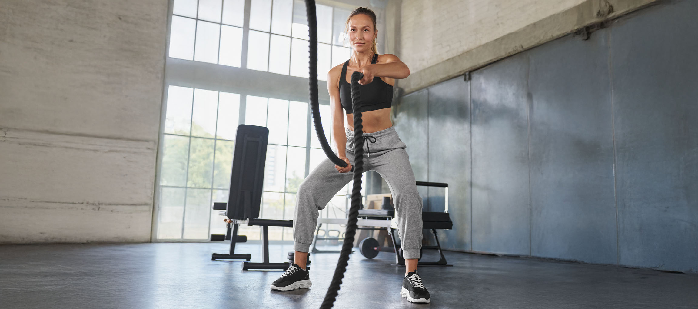 Vrouw in zwarte sportbeha en grijze joggingbroek oefent met battle ropes