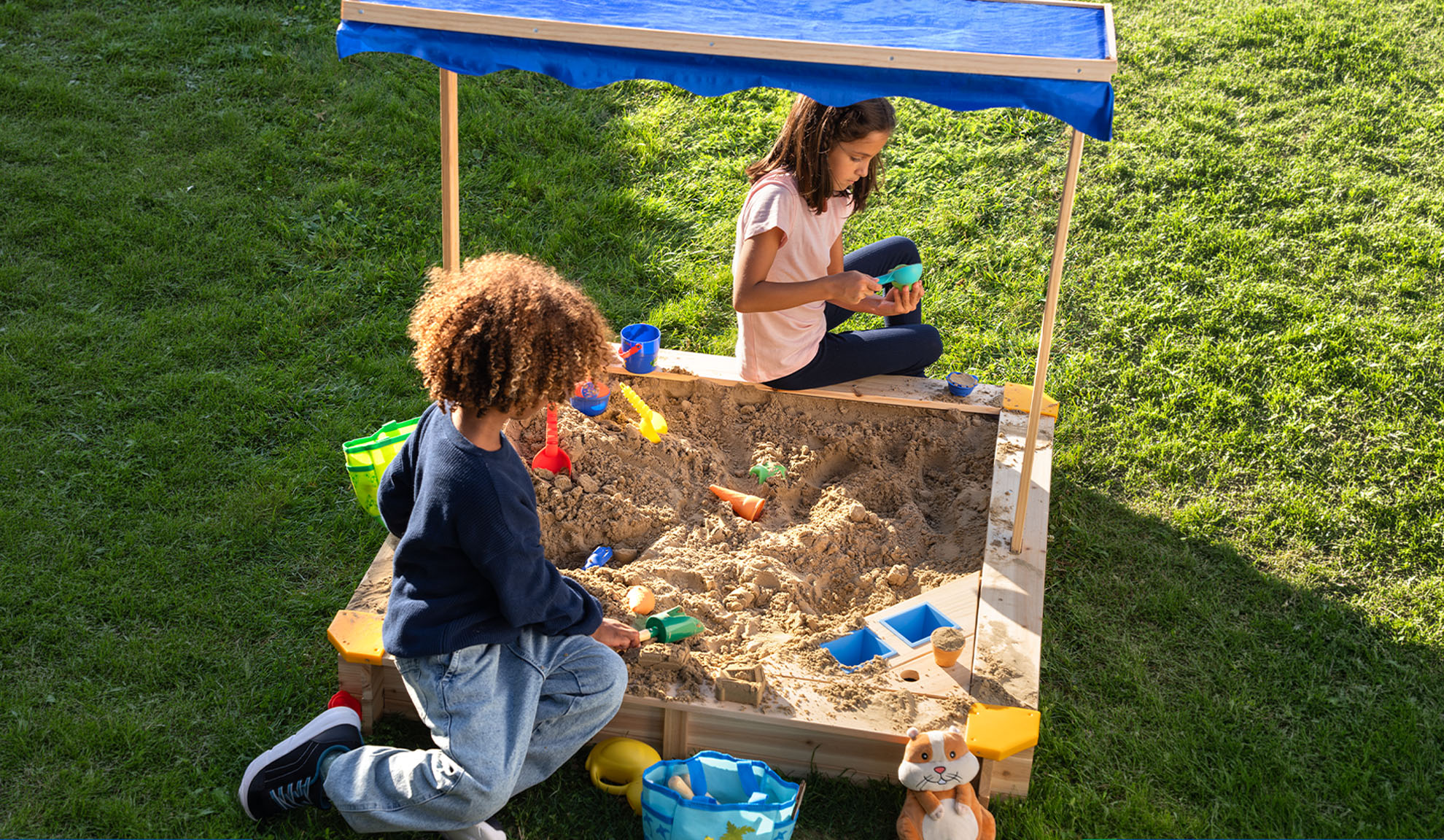 Twee kinderen spelen in een zandbak met blauwe luifel en zandspeelgoed.