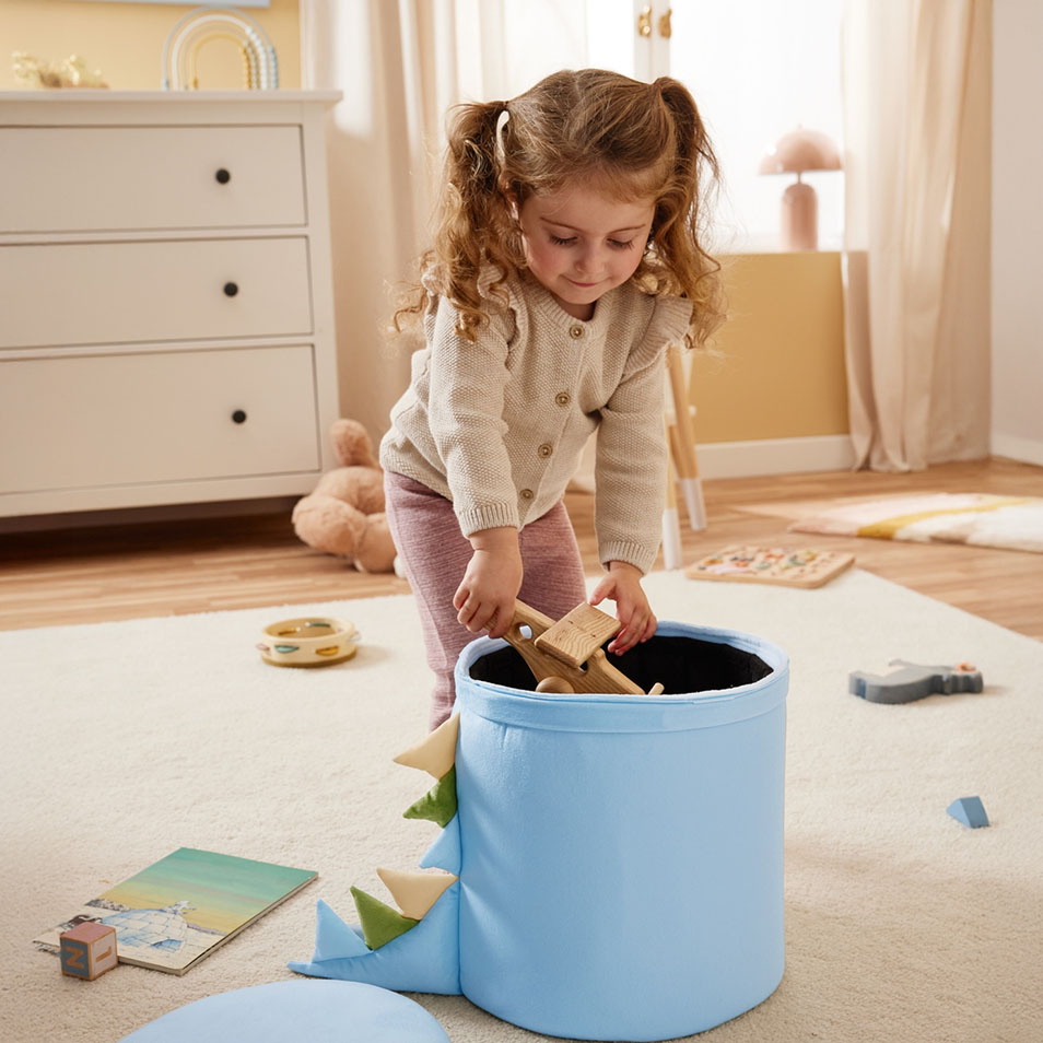 Een kind stopt houten speelgoed in een blauwe opbergbak met dinosaurus-thema in een slaapkamer.