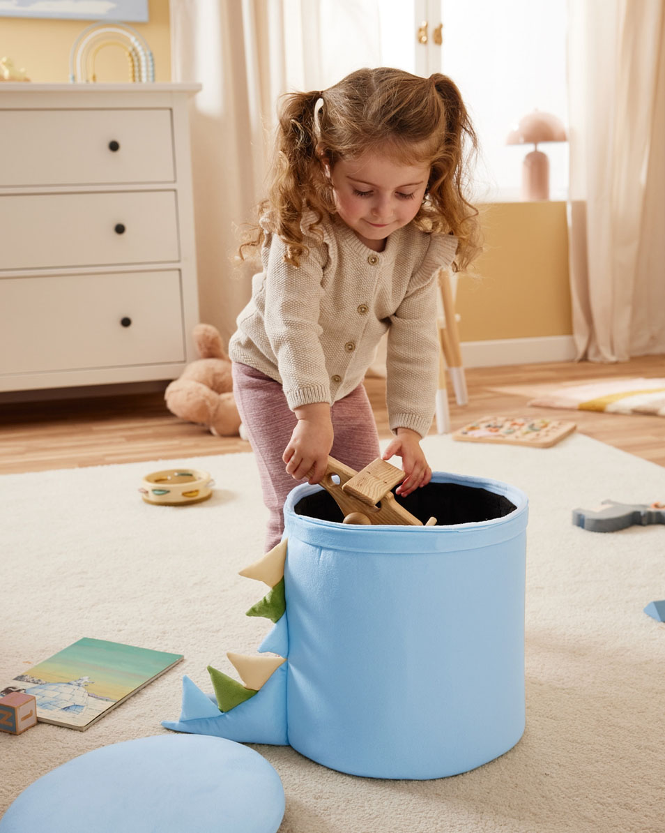 Een kind stopt houten speelgoed in een blauwe opbergbak met dinosaurus-thema.