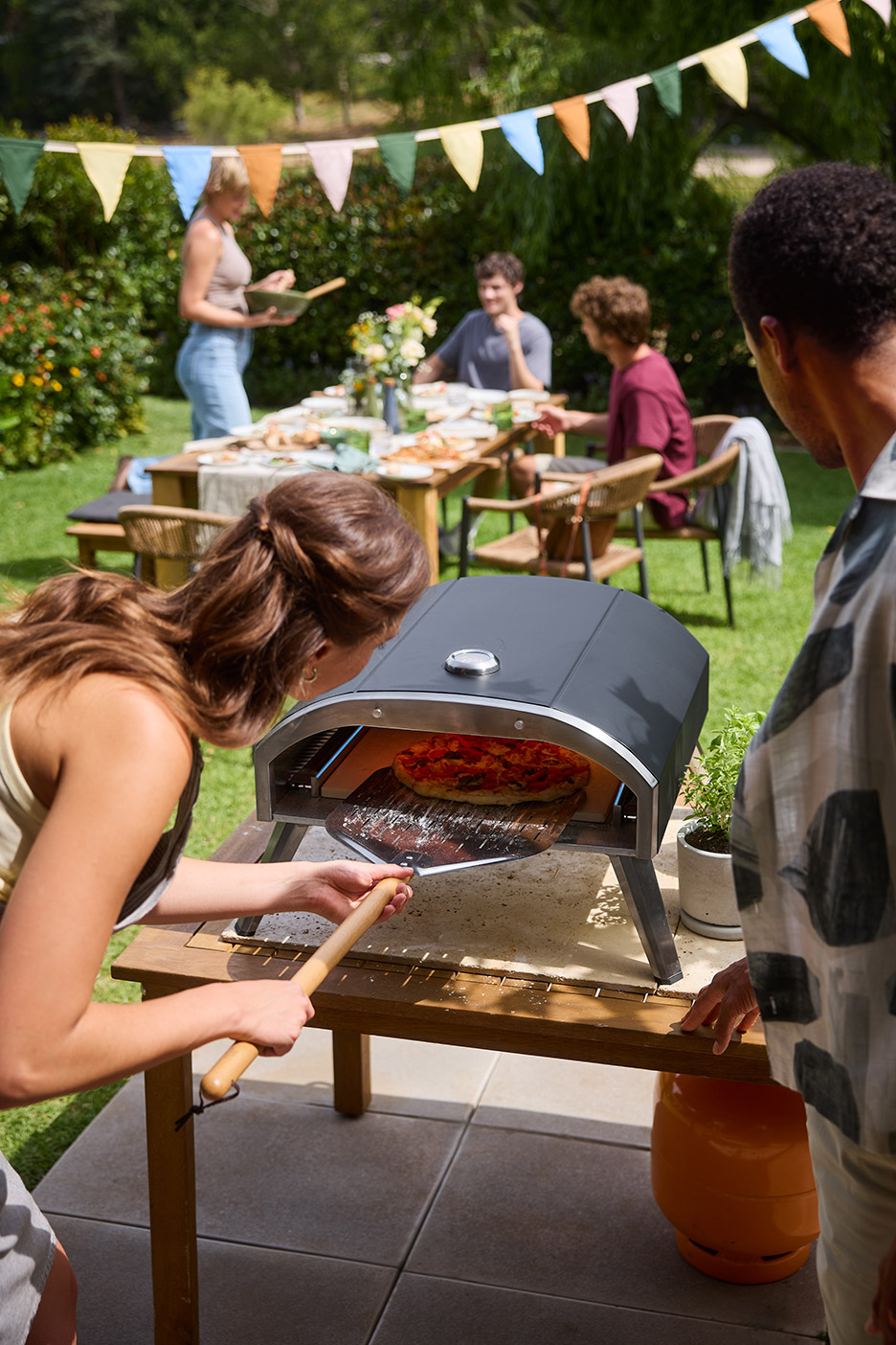 Vrouw haalt pizza uit een pizzaoven op een tuintafel, met vrienden op de achtergrond.