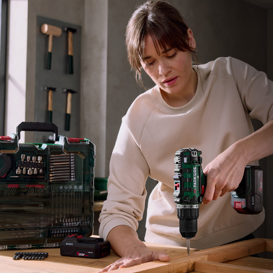 Vrouw boort in hout met een accuboormachine, naast een gereedschapskist en een accu.