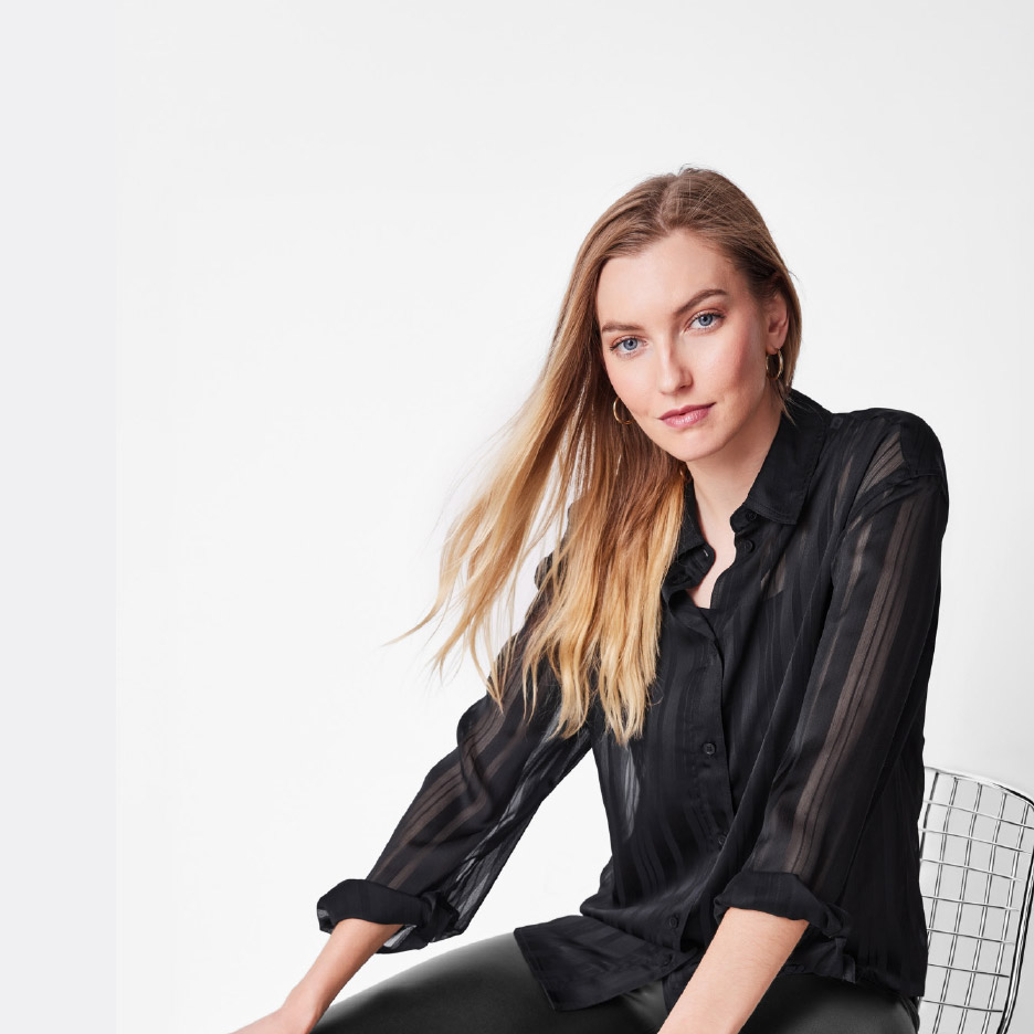 Vrouw in een zwarte, doorschijnende blouse en zwarte broek, zittend op een stoel.