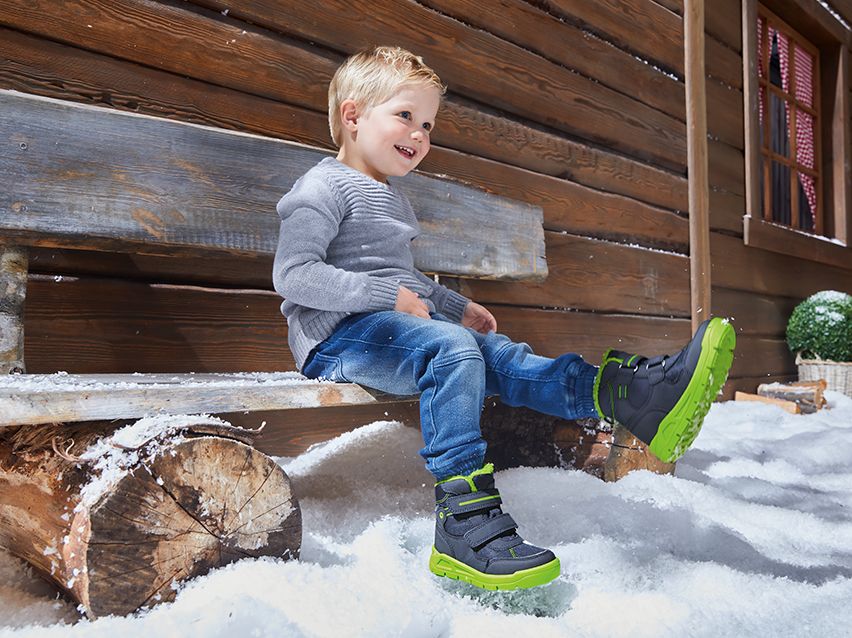 Lachende jongen in grijze trui, jeans en donkerblauwe en limoengroene winterlaarzen in de sneeuw.