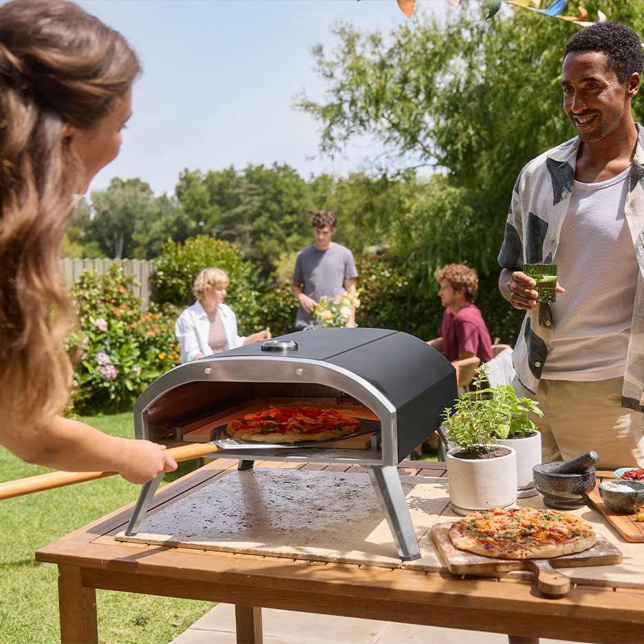 Familie op een tuinfeest, vrouw stopt pizza in een oven, man houdt een drankje vast.